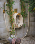 Hose hook in raw brass mounted on a wall with garden hose and faucet