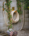 Hose hook in raw brass mounted on a wall with garden hose and faucet